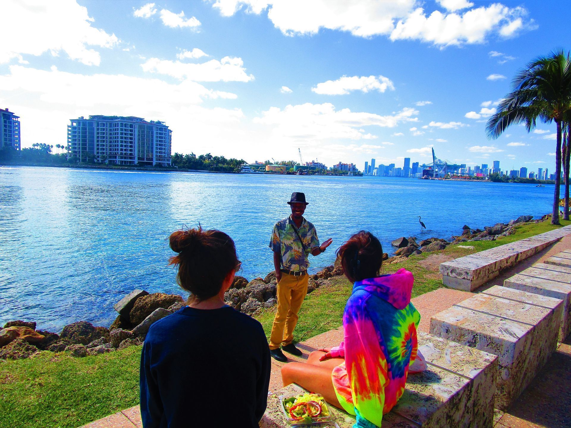 People by a waterfront, one playing guitar, with blue water, skyline, and palm trees under a bright sky