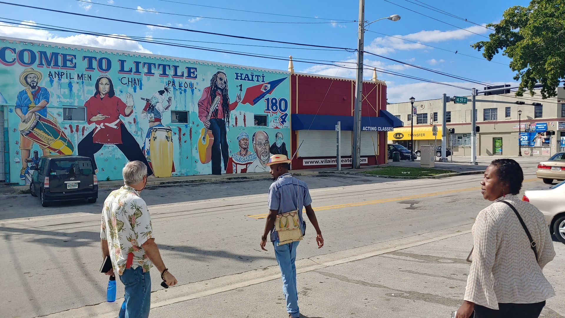 People crossing a street beside a colorful mural and a red building on a sunny day