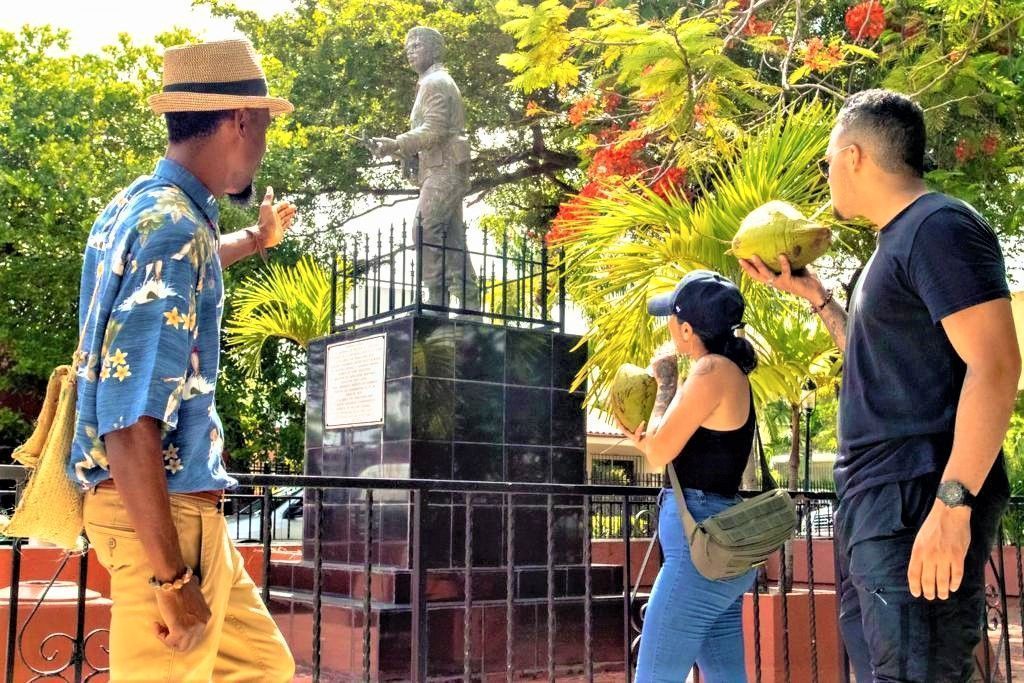 Three people looking at a statue in a sunny park with red flowers and a fence.