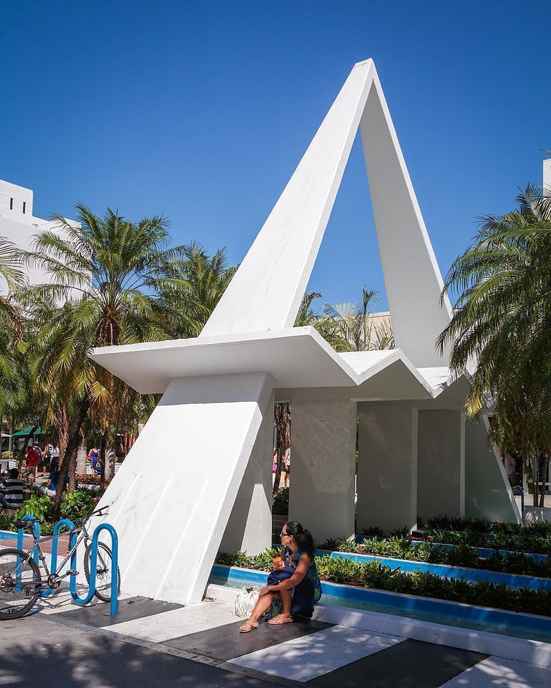 White modern sculpture with triangular beams in a sunny plaza, with palm trees and a person sitting nearby