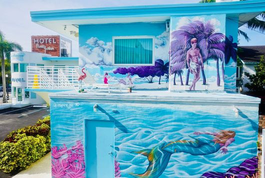 Colorful ocean mural on a building, with swimmers, tropical flowers, and a hotel sign in the background