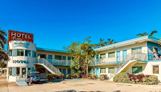 Sunny exterior of a two-story hotel with white walls, blue accents, stairs, and a sign by the entrance