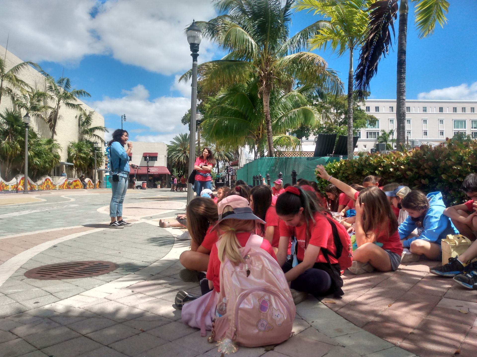 Children sitting on a sunny plaza, listening to a speaker near palm trees and a white building.