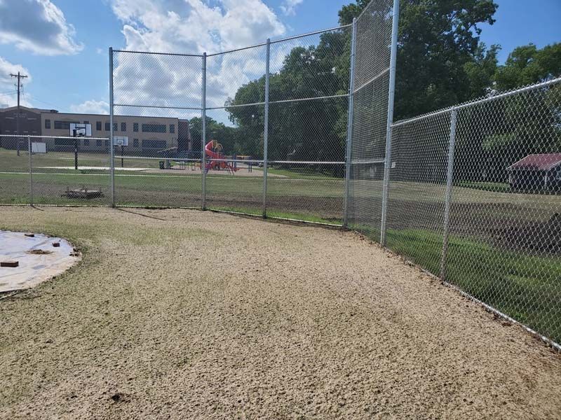 Chain-link fenced baseball field with gravel foreground and playground equipment in the distance.