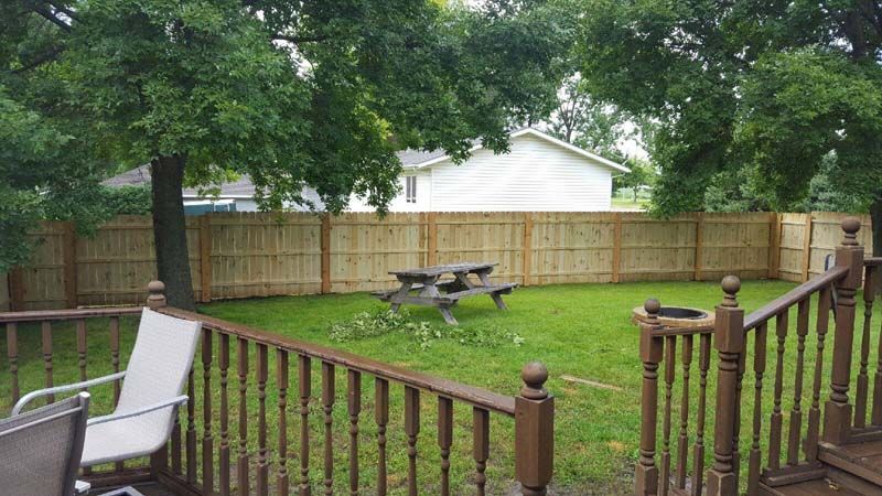 Backyard with wooden fence, picnic table, and deck railing under leafy trees after rain