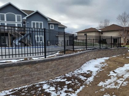 Snowy suburban backyard with stone retaining wall, black fence, and two houses under overcast sky