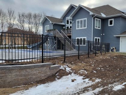 Blue-gray house with a black metal fence and snowy yard in a wooded suburban setting
