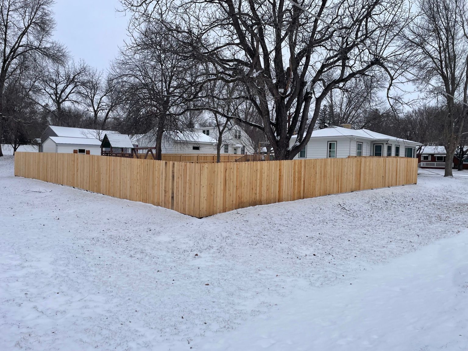 Snow-covered yard with a new wooden privacy fence and bare trees in the background