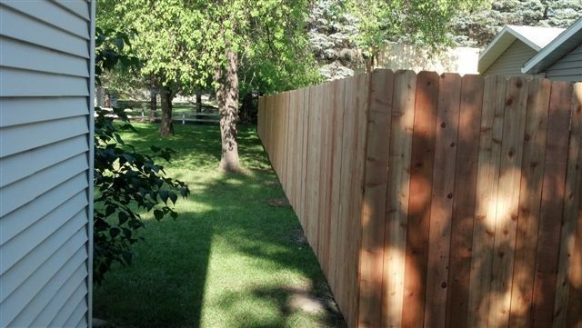 Narrow grassy side yard between a house and a wooden privacy fence, with trees in the background