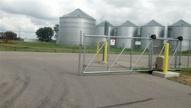 Farm grain silos behind a closed chain-link gate on a paved lot under cloudy skies
