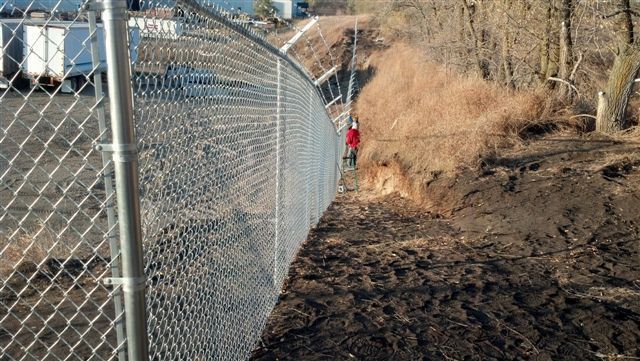 Chain-link fence along a muddy path with a person in red in the distance near dry brush.