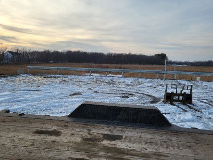 Snow-covered field with a small ramp, dirt foreground, and a bench-like structure under a cloudy sky