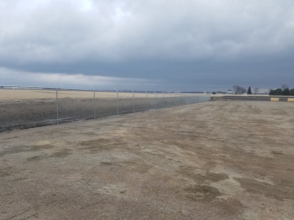 Cloudy sky over an empty gravel lot beside a chain-link fence and open field
