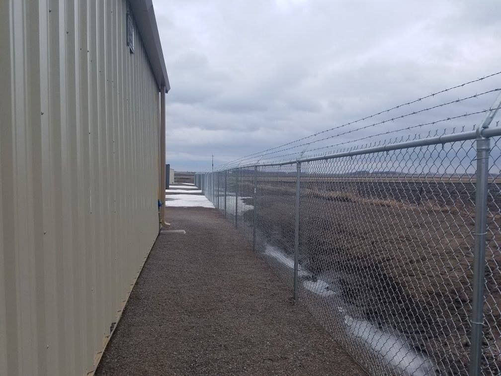 Narrow gravel path beside a tan building and chain-link fence under a cloudy sky
