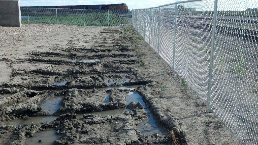 Muddy excavation site beside a chain-link fence, with standing water and a grassy embankment.