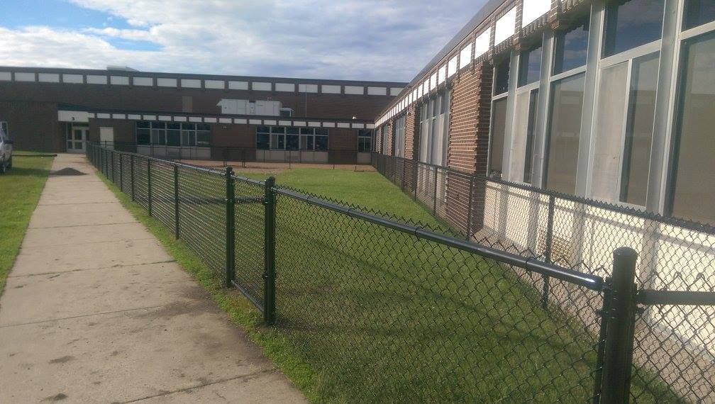 School courtyard with a concrete path, grassy lawn, chain-link fence, and long brick-and-glass building.