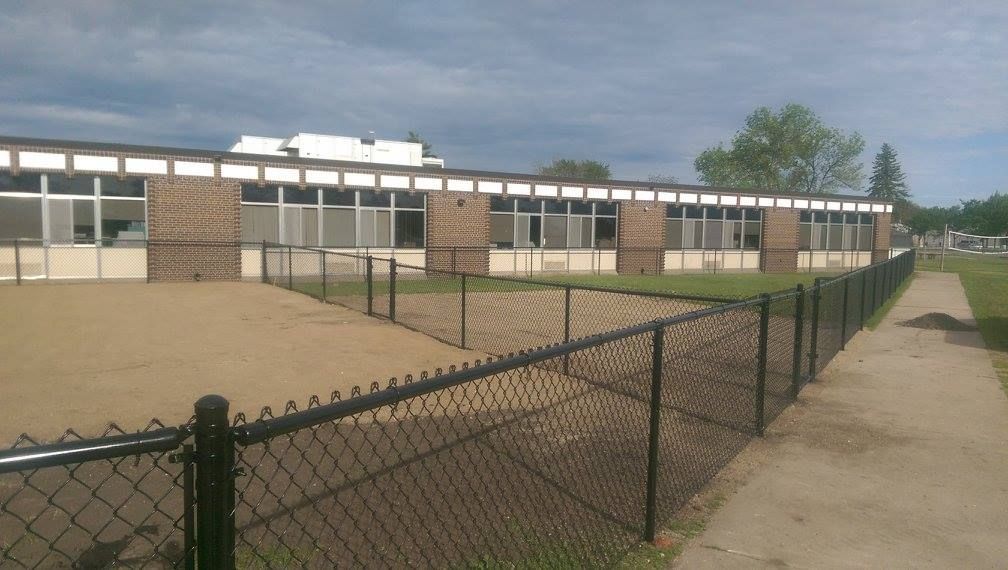 School courtyard with single-story classrooms, chain-link fence, and sandy playground under a cloudy sky