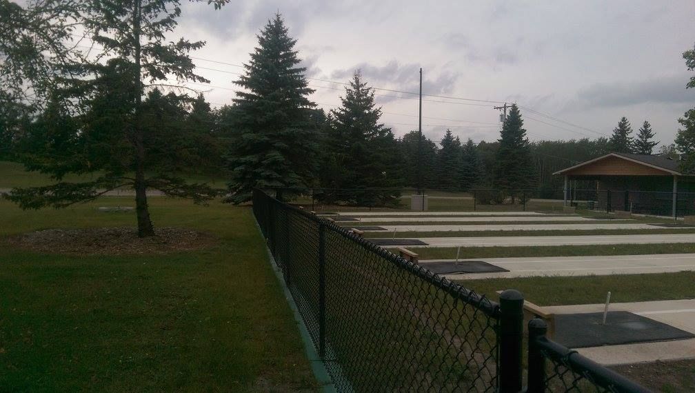 Chain-link fence beside a grassy park, with evergreen trees and a picnic shelter under cloudy skies.