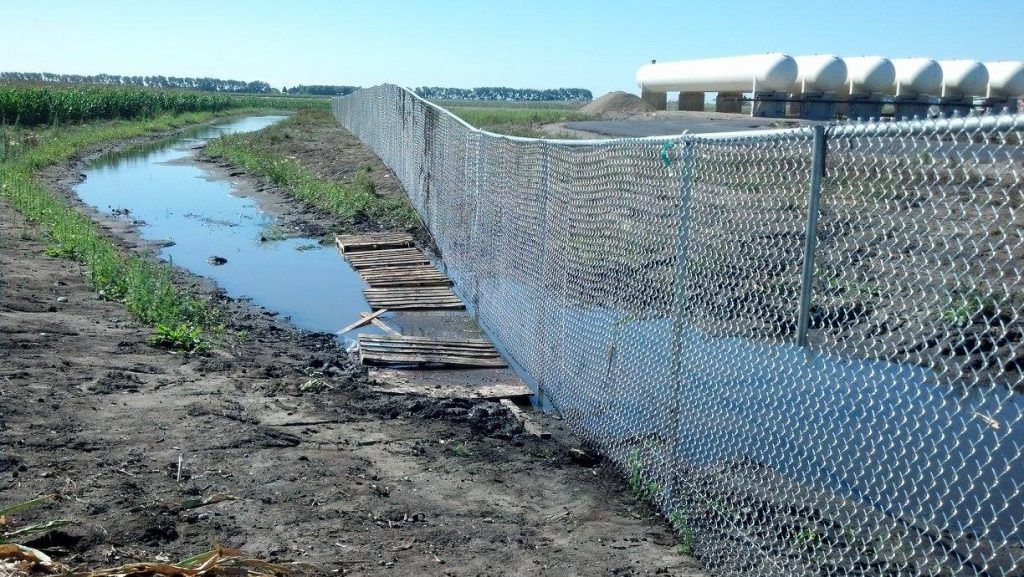 Chain-link fence beside a narrow water canal in a rural field under a clear sky