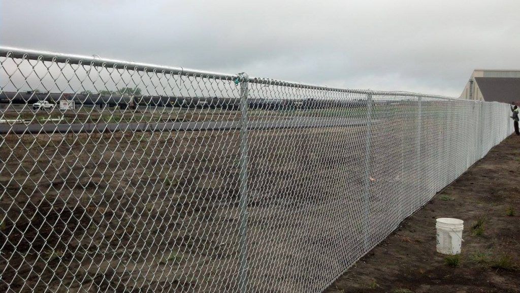 Chain-link fence along a muddy roadside beside open fields under a cloudy sky.