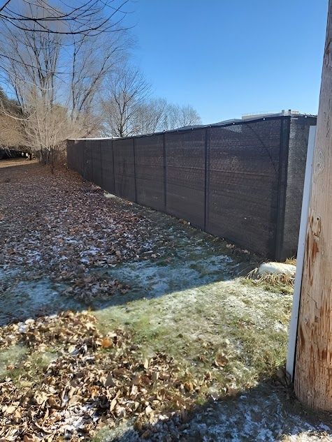 Snow-dusted backyard with a long dark fence, bare trees, and fallen leaves on the ground.