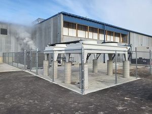Industrial facility with fenced metal equipment on a concrete pad beside a large gray warehouse