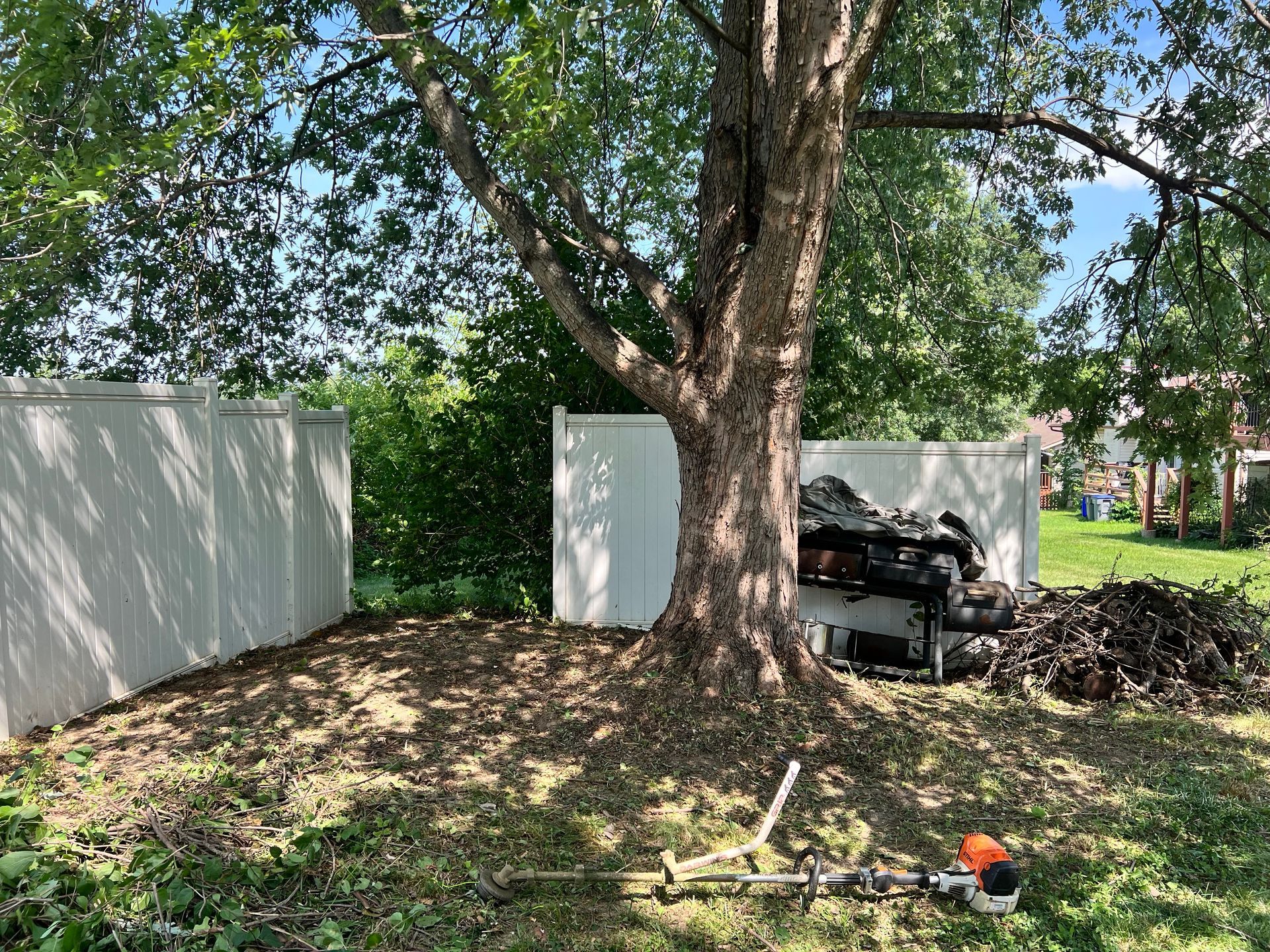 A lawn mower is sitting under a tree in a yard next to a white fence.