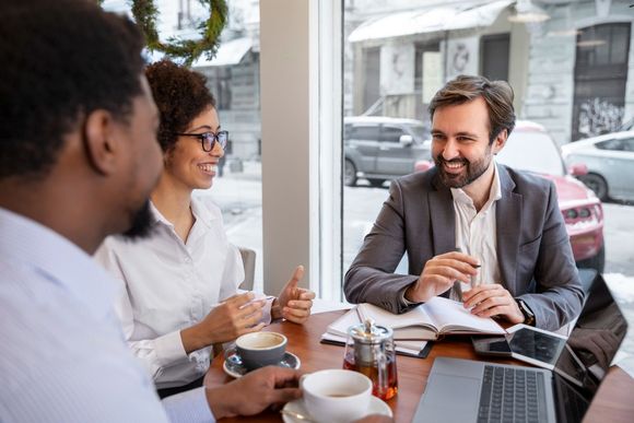 Three people in business attire at a cafe table, smiling and conversing; laptop and coffee present.
