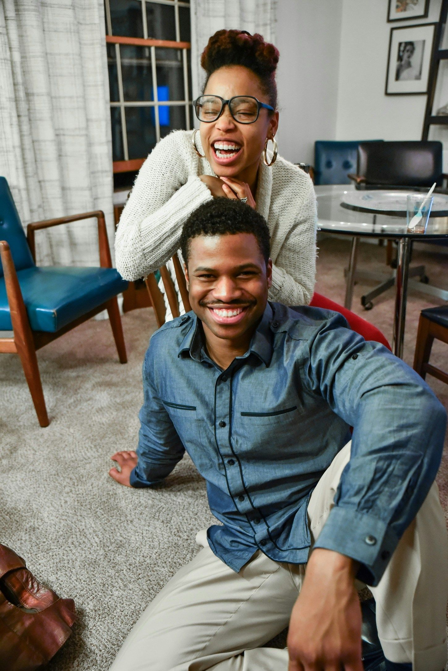 Smiling person on a rug with another person behind, laughing. Living room setting with furniture and a window.