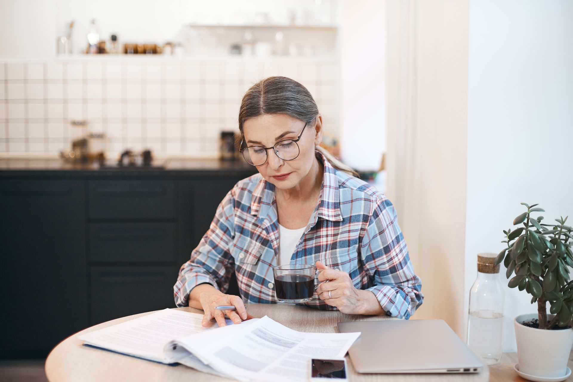 Woman with glasses, plaid shirt, looks at paperwork, sitting at a table in a kitchen.