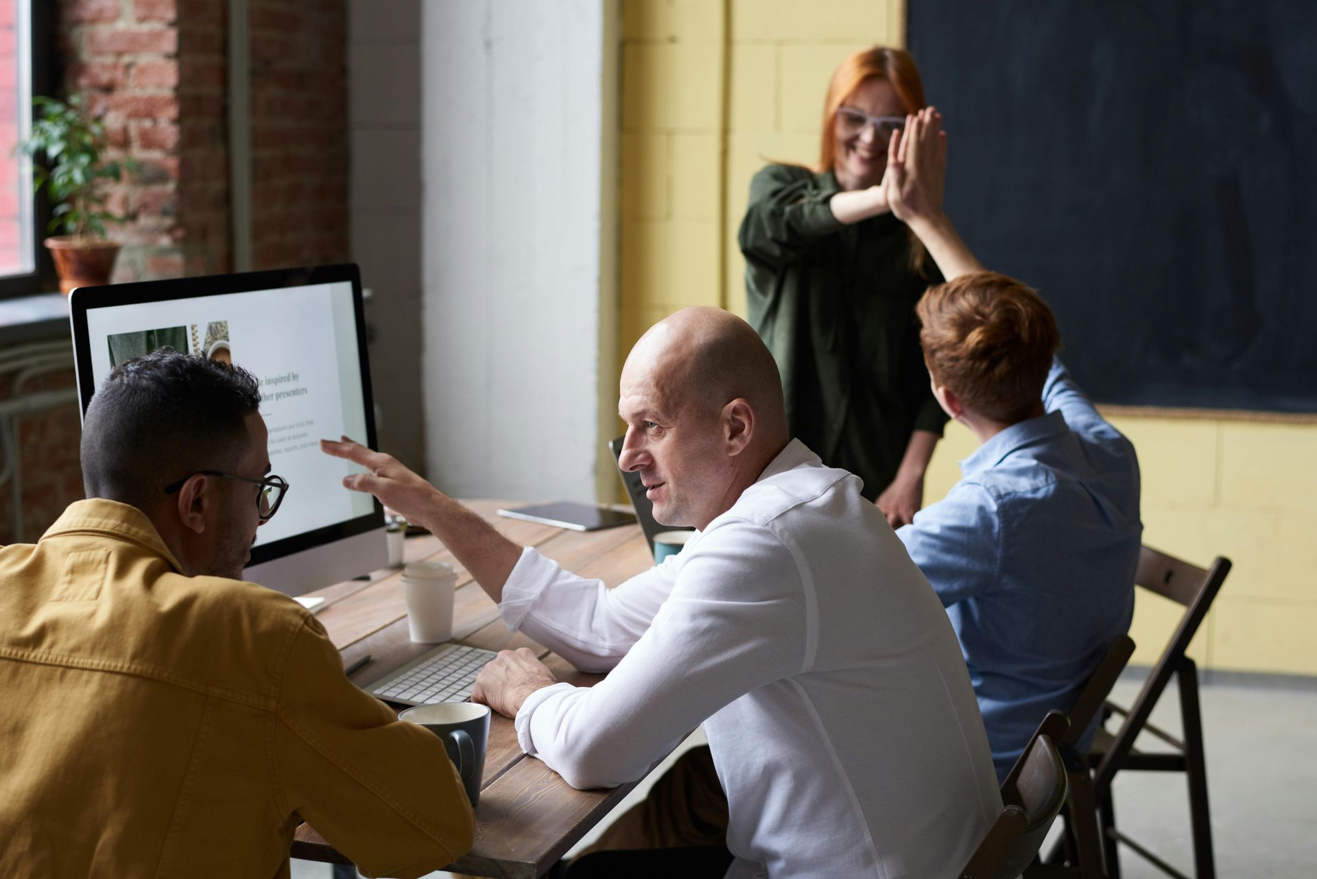 Team meeting around a computer. Two men at the table, others in the background giving high-fives.