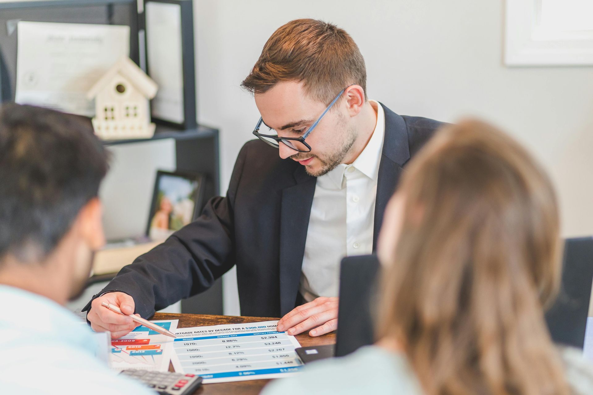 A man in glasses points at paperwork, discussing with a couple at a table.