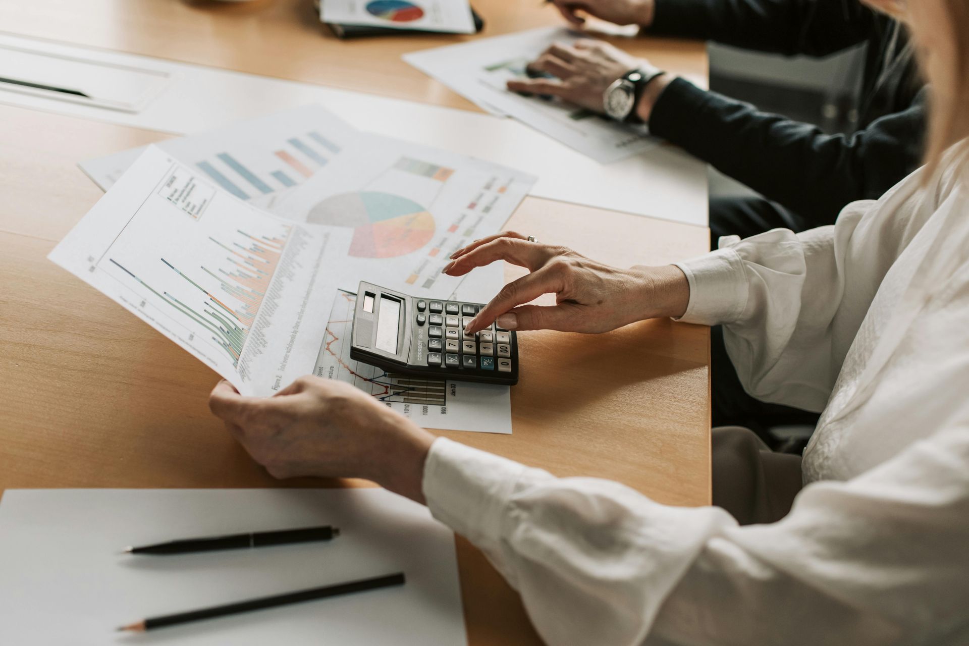 Person using a calculator, reviewing financial reports with graphs, at a table with others.