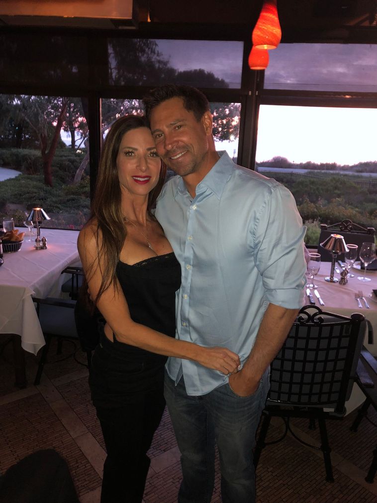 Couple posing together at a waterfront restaurant during sunset, smiling and dressed for dinner.
