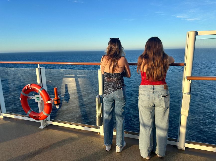 Two people in jeans stand by a boat railing, looking out over the ocean at sunset.
