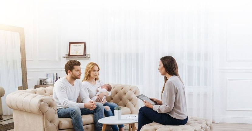 Three people talking in a bright living room, seated on a sofa and chair around a small table.