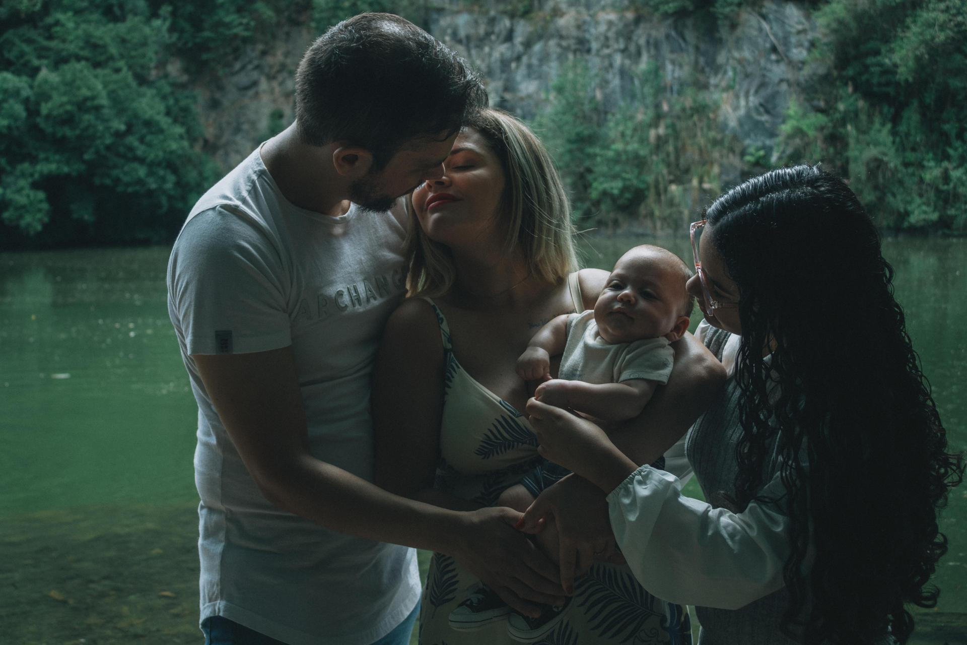 Family holding a baby by a forest river, with greenery reflected in calm water.