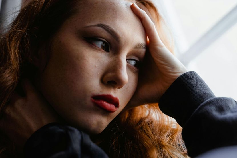 Thoughtful woman with red hair resting by a window, looking away with her hand on her forehead