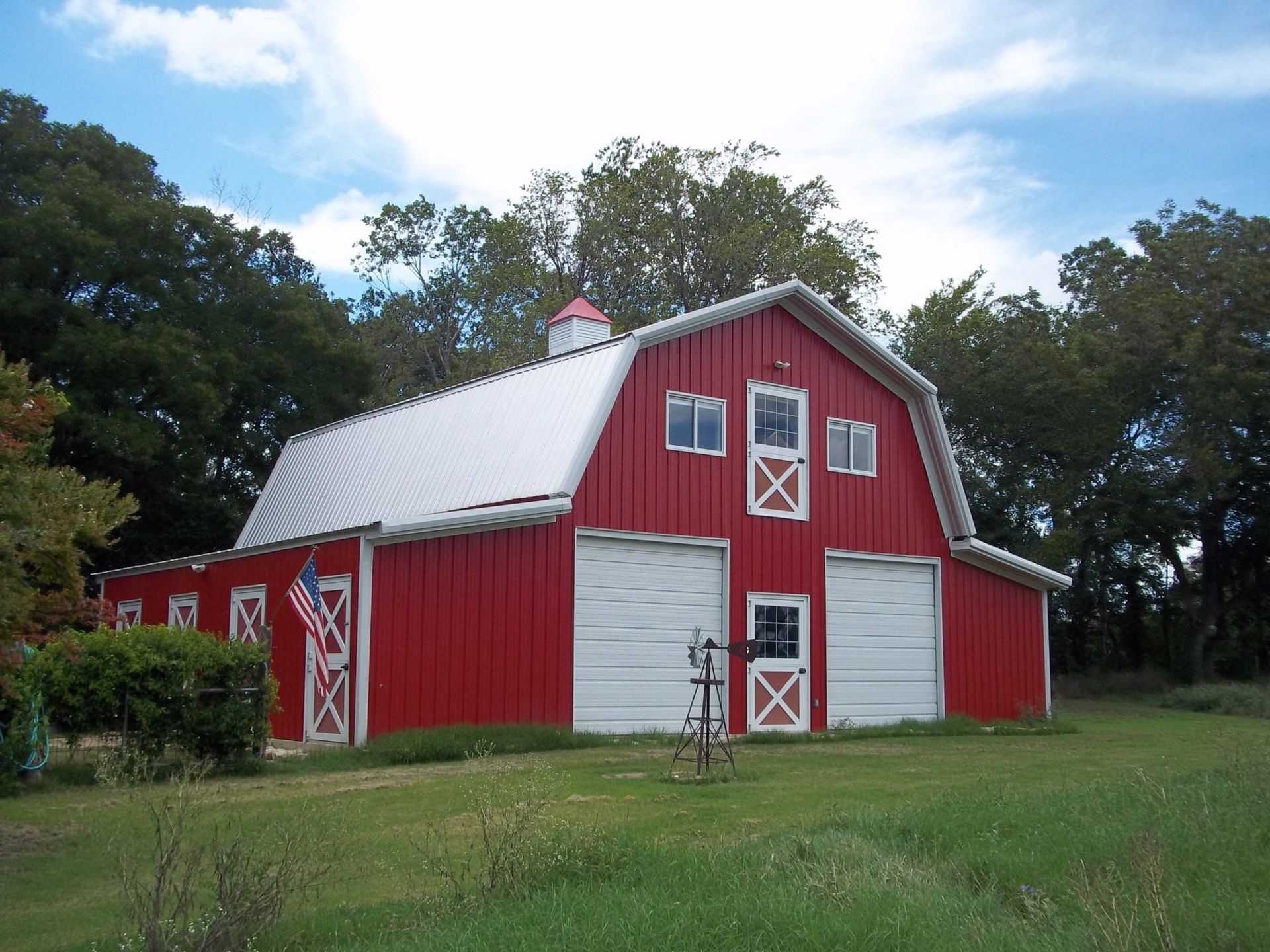 Red barn with white doors and roof, American flag, set in a grassy field with trees and blue sky.