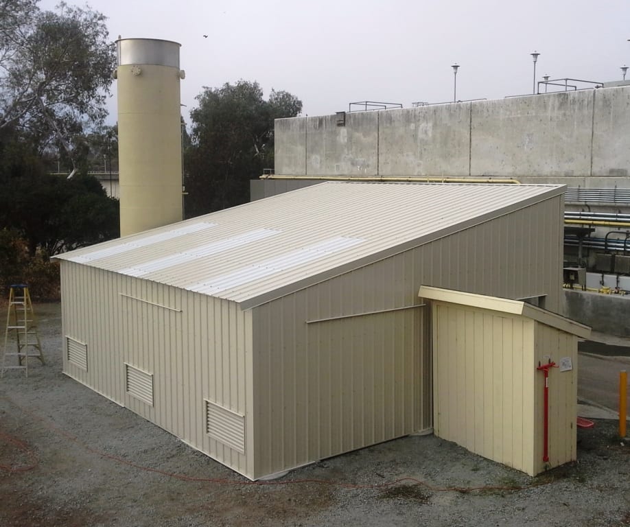 Tan metal building with a shed extension, near a tall silo and other industrial structures.