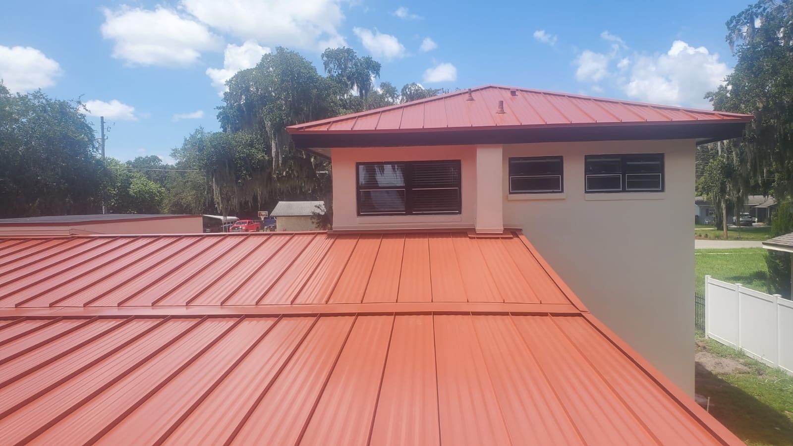 Red metal roof of a building with a square structure and windows. Blue sky and trees are in the background.
