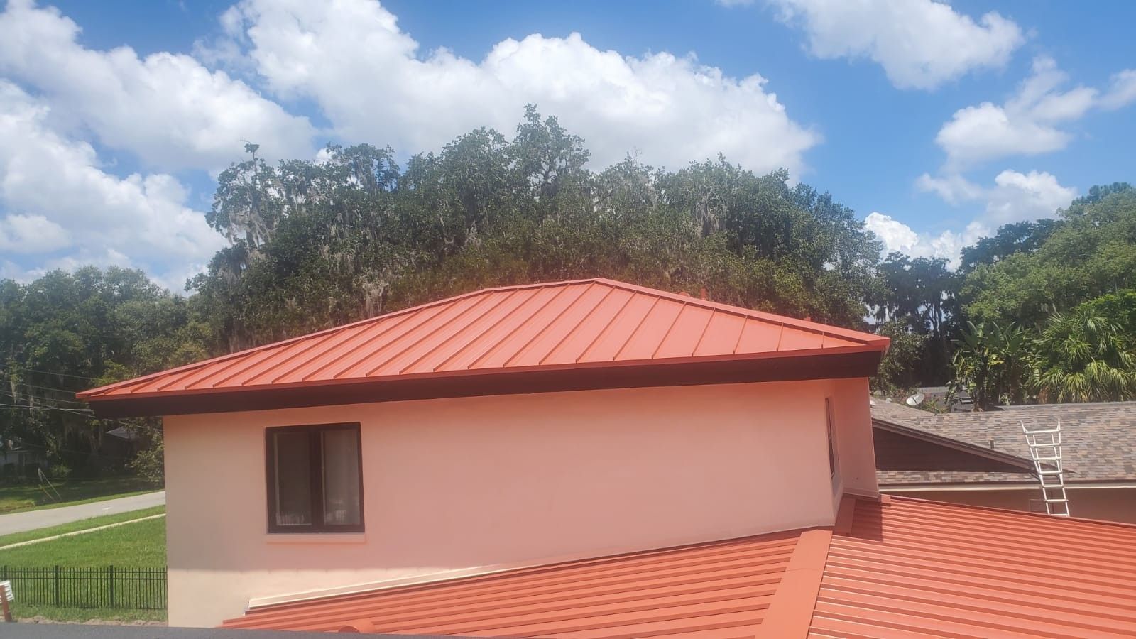Red metal roof on a light-colored house against a cloudy blue sky. Trees in the background.