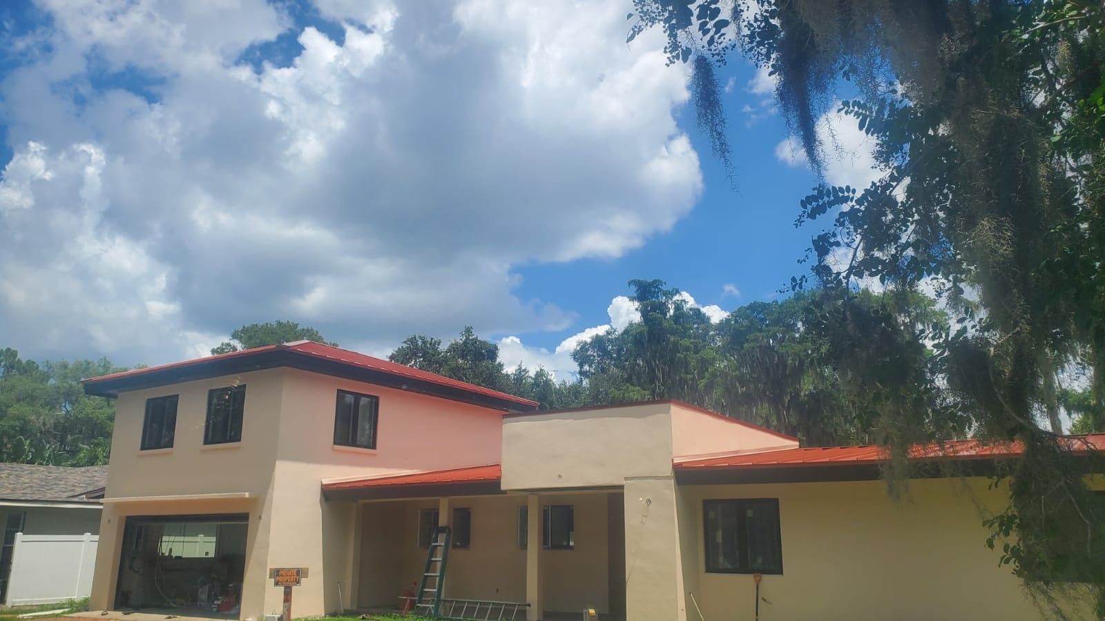 Two-story tan and peach house with red-tiled roof under a cloudy blue sky, with trees.