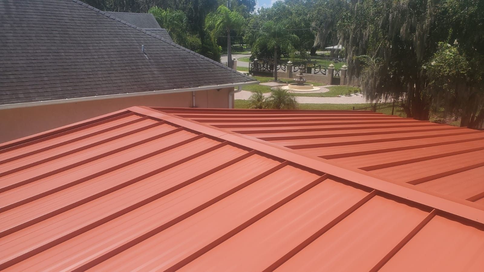 Red metal roof on a house, with a gray shingle roof in the background and a view of trees and homes.