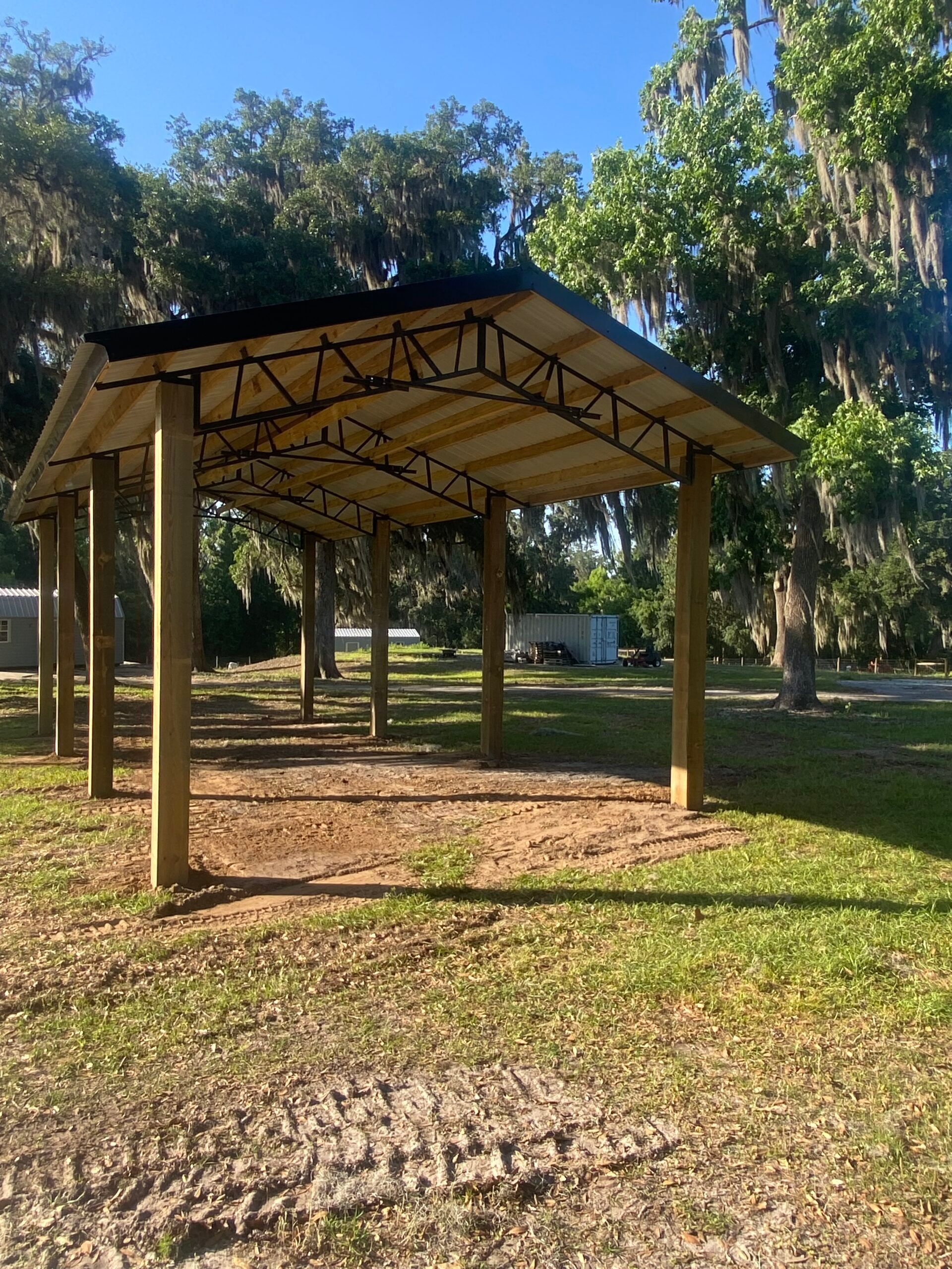 Wooden carport in a grassy area, surrounded by trees under a blue sky.