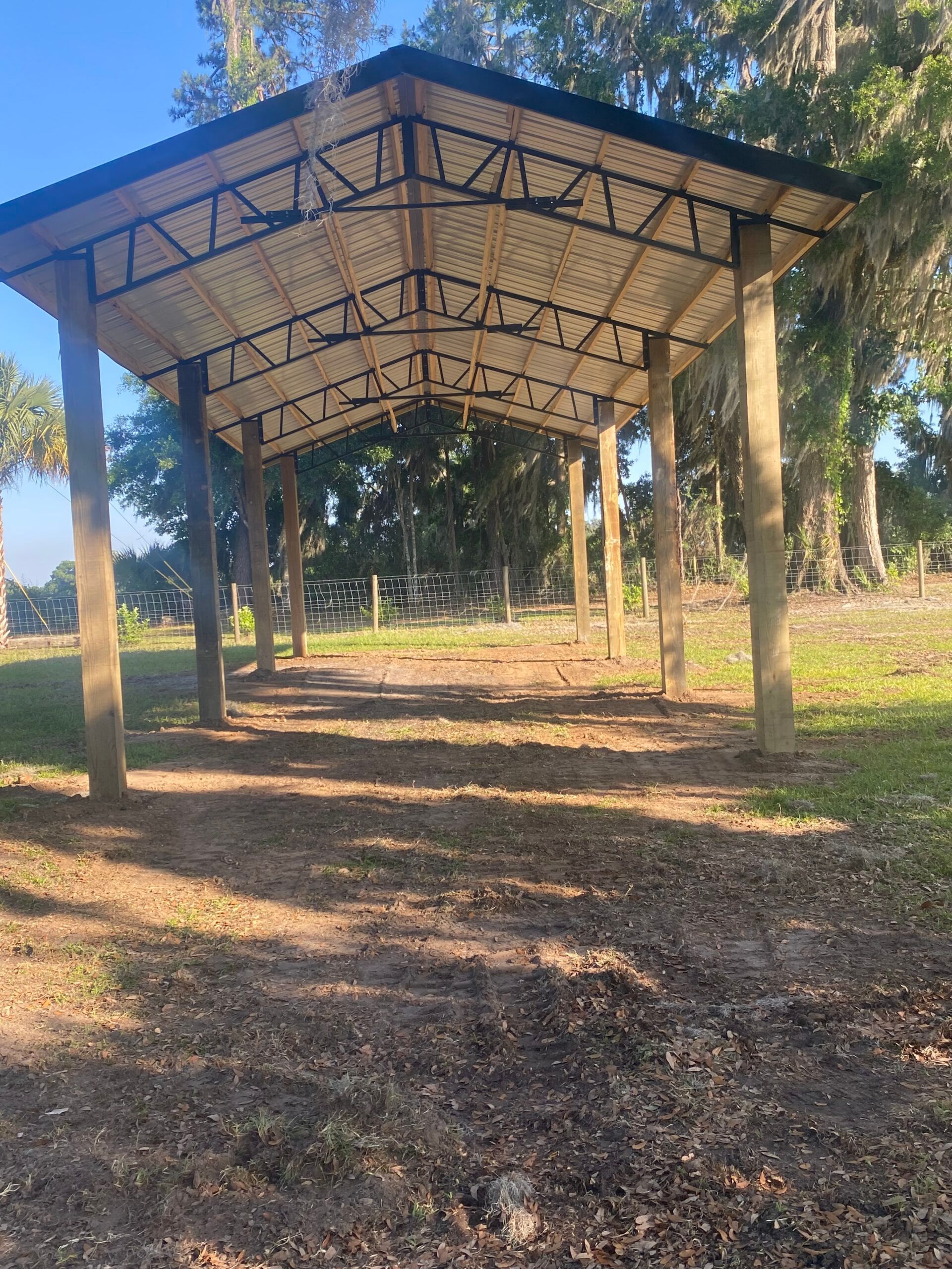 Wooden shelter with posts, corrugated roof, dirt floor, trees in background.