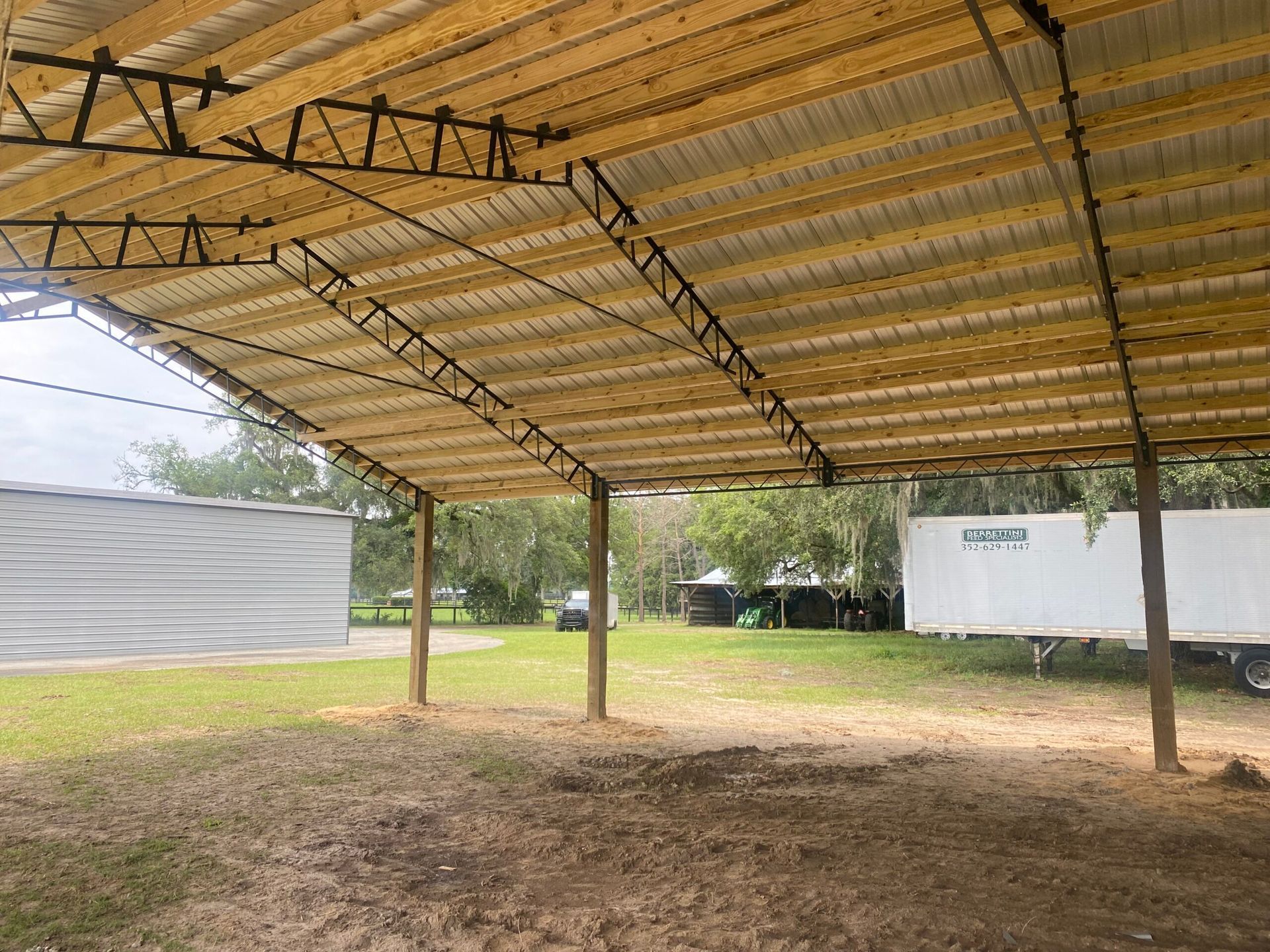 Open-air wooden structure with a corrugated metal roof, supported by posts, in a grassy area.