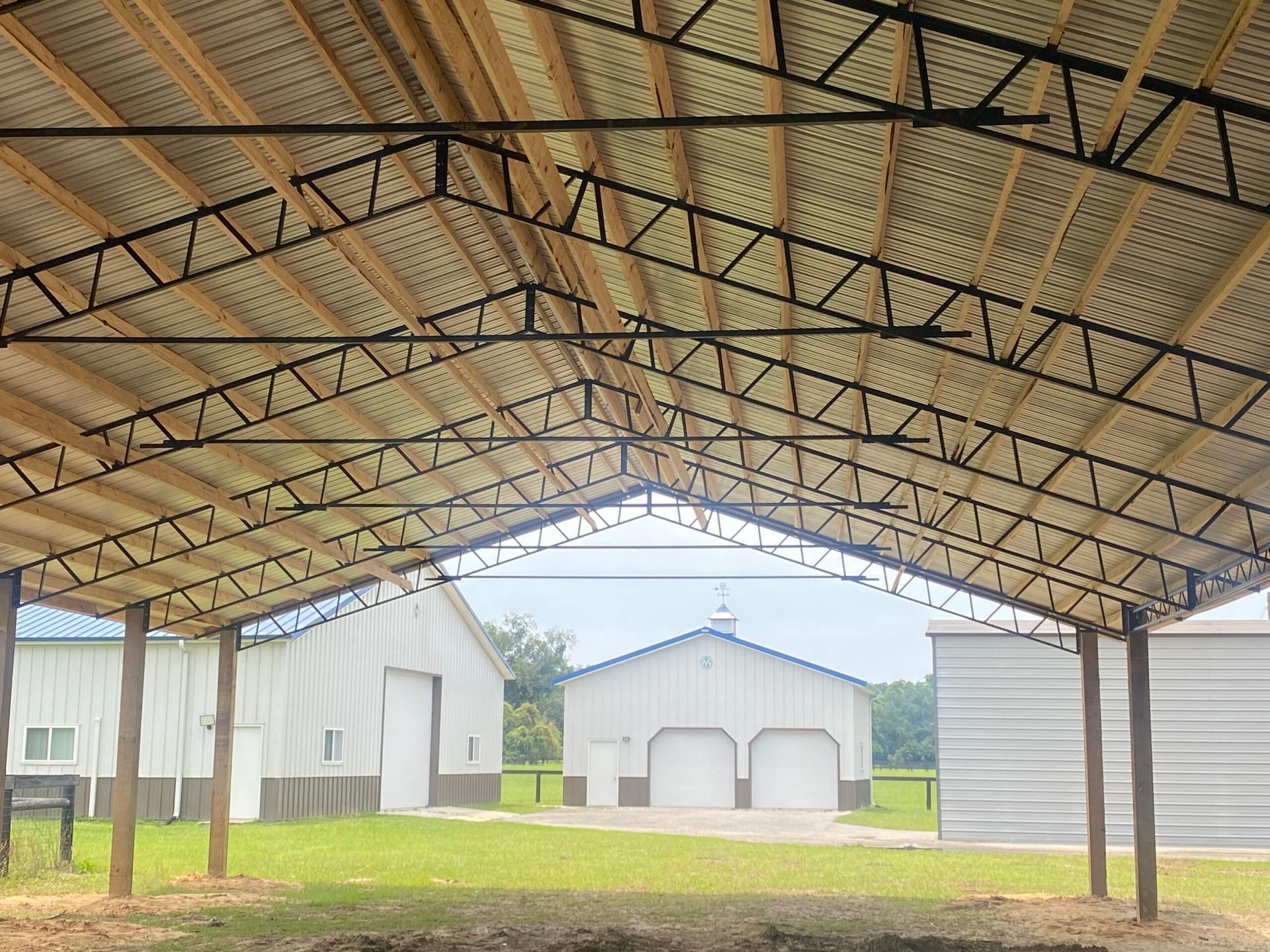 Interior view of a large metal and wood structure with a view of several white farm buildings and a green field.