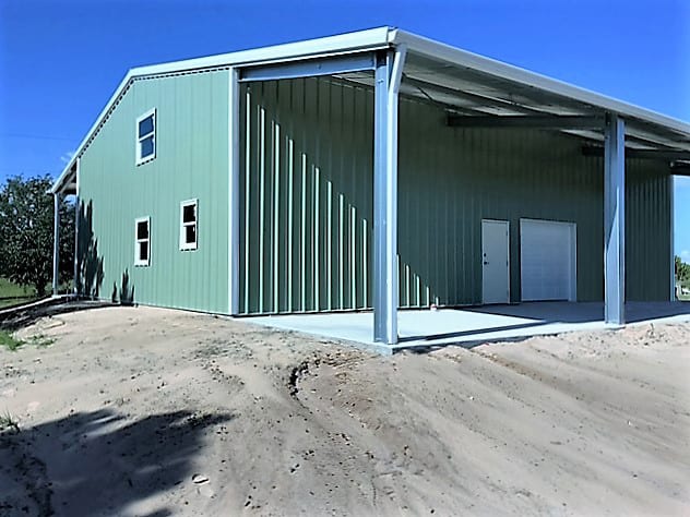Green metal building with an open-air awning, a concrete pad, and small windows.