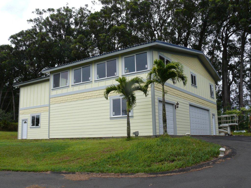 Two-story light yellow house with large windows, palm trees, and a garage in a grassy setting.